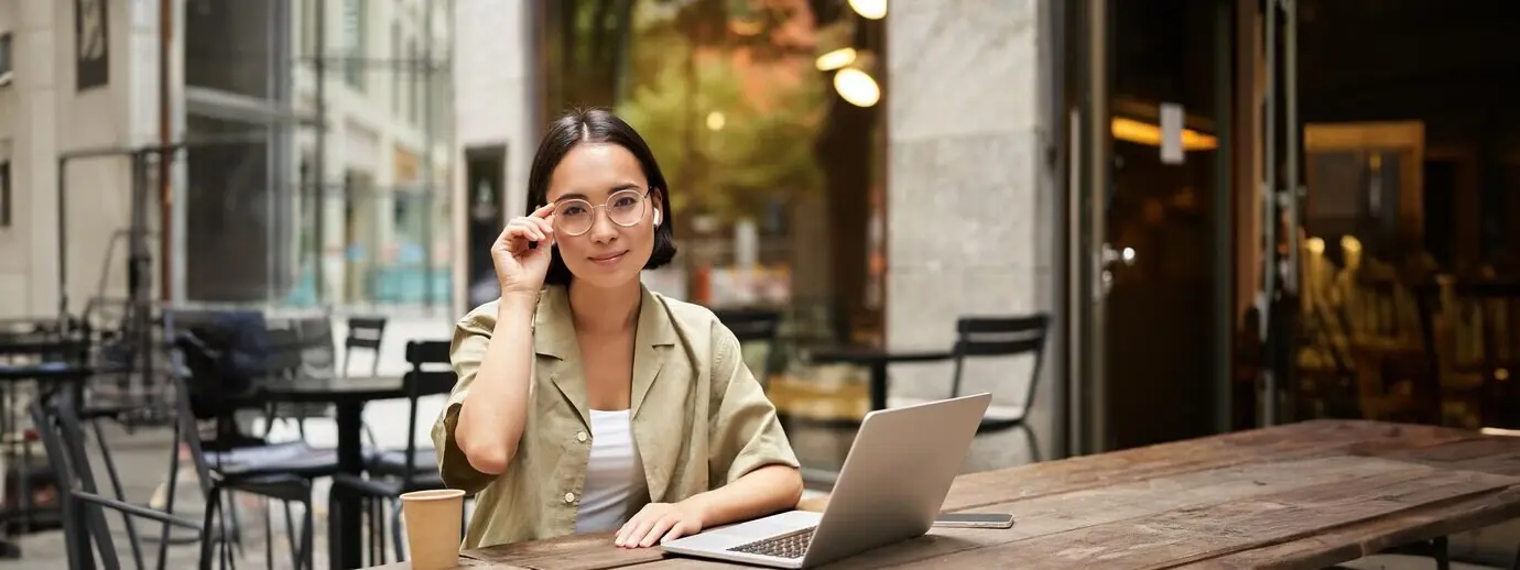 Молодая студентка работает из кафе, сидя на открытом воздухе с ноутбуком и чашкой кофе и смотрит.